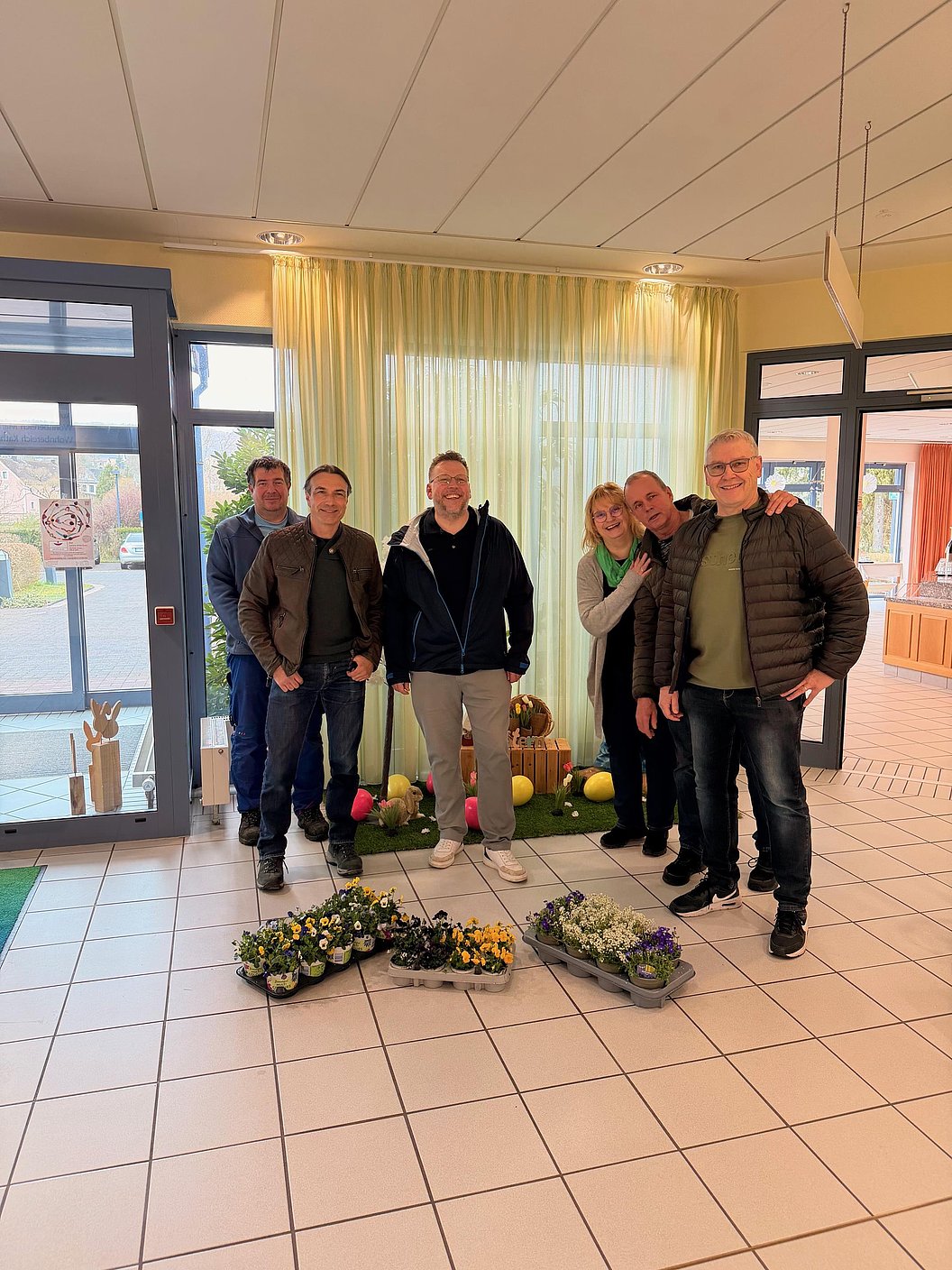 Sechs Personen stehen in einem hellen Raum mit großen Fenstern hinter mehreren auf dem Boden arrangierten Blumensträußen
