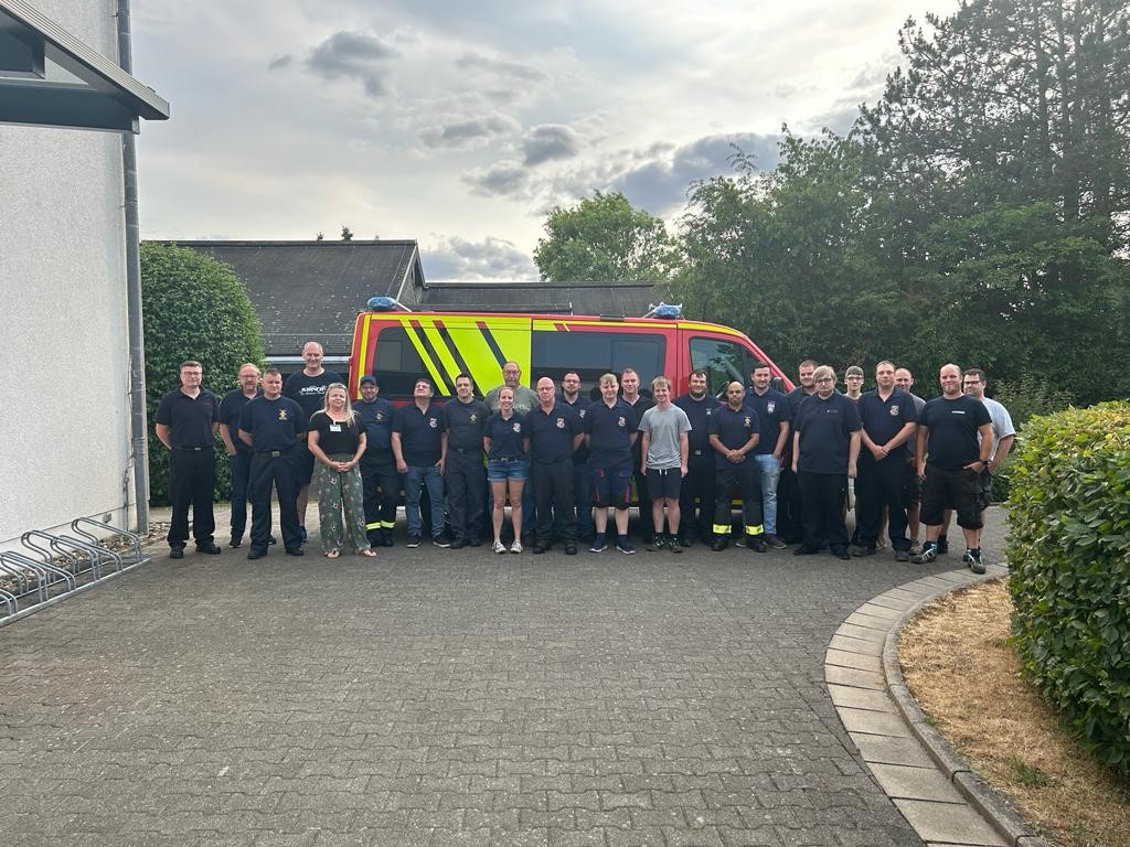 Gruppe von Feuerwehrleuten in Uniform vor einem roten Feuerwehrfahrzeug, das an einem Gebäude und Bäumen geparkt ist.