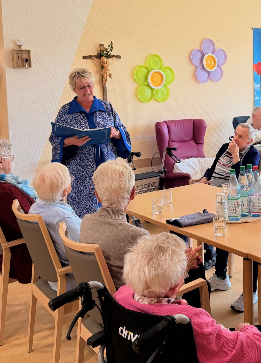 Ältere Menschen sitzen an einem Tisch und hören einer stehenden Frau zu, die ein Buch hält, im Raum mit Wanddekoration aus Blumen und einem Kreuz